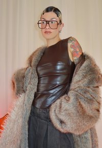 Haut marron sans manches en cuir sous un manteau en fausse fourrure, avec une jupe en denim gris. Grandes lunettes épaisses et tatouage floral coloré visible.