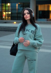 Mint grønn Nike hettegenser og matchende joggebukser med brodert logo. Modellen har på seg hodetelefoner og holder en svart veske. Urban bakgrunn.