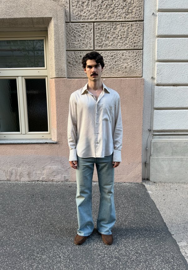 Junger Mann mit dunklem Haar und Schnurrbart, der ein hell gestreiftes Hemd, blaue Jeans und braune Schuhe trägt, steht an einer strukturierten urbanen Wand in der Nähe eines Fensters.
