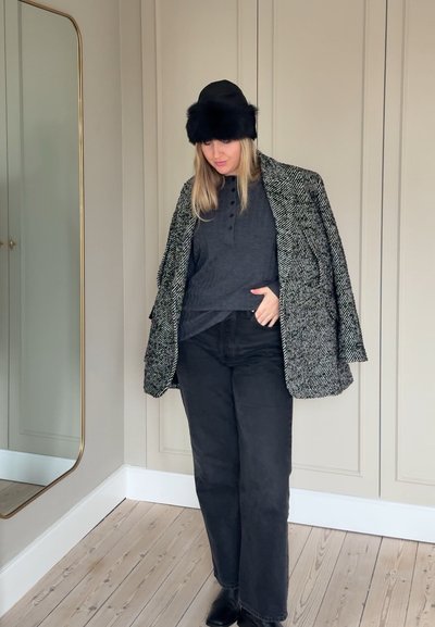 Blazer de espiga en blanco y negro sobre una camiseta henley gris oscuro, combinado con pantalones de pierna ancha negros y un sombrero de piel negro; de pie en una habitación con suelo de madera clara.