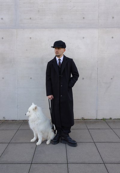 Abrigo largo negro sobre un traje negro, camisa blanca y corbata. Un perro Samoyedo blanco se sienta al lado de la persona, que lleva botas negras con suelas gruesas.