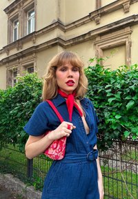 Denim jumpsuit i mørkeblå, med korte ærmer, et bælte med en rund spænde og en dyb udskæring. Accentueret med et rødt tørklæde og taske.
