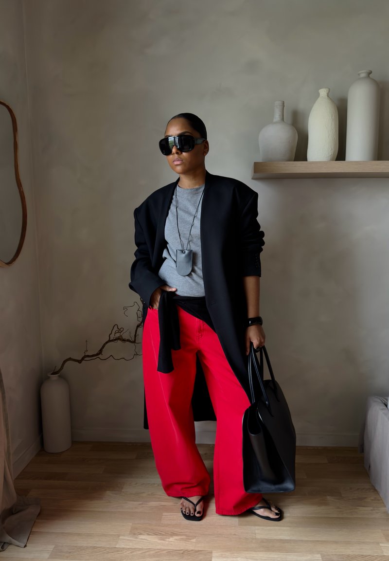 Femme portant des grandes lunettes de soleil noires, un blazer noir, un haut gris, un pantalon rouge à jambes larges, tenant un sac cabas noir dans une pièce minimaliste avec des vases.