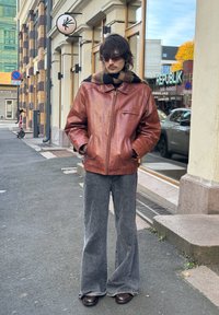 Homme portant une veste en cuir marron, une écharpe rayée noire et marron, des lunettes de soleil et un pantalon gris à jambes larges, debout sur un trottoir urbain près d'une vitrine.