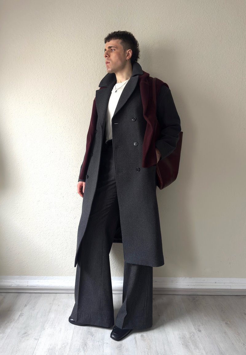 Persona in piedi al chiuso che indossa un lungo cappotto grigio scuro, pantaloni a gamba larga, camicia bianca, maglione bordeaux drappeggiato sulle spalle e una borsa tote bordeaux.