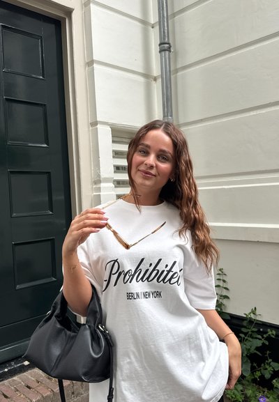 Camiseta blanca de gran tamaño con el texto "Prohibité" y "BERLIN | NEW YORK", bolso de cuero negro y gafas de sol sostenidas en una mano.