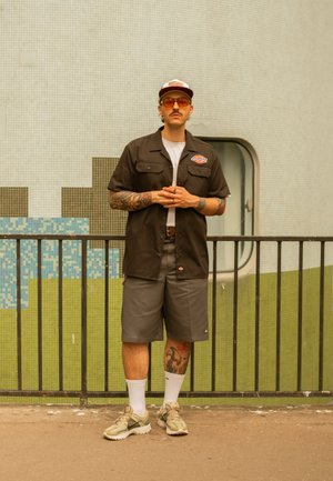 Schwarzes kurzärmliges Shirt, graue Shorts, darunter ein weißes T-Shirt. Braune Kappe, orange getönte Sonnenbrille, Sneaker mit hellgrünen Akzenten.