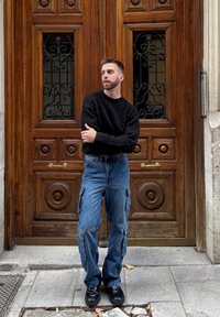 Suéter preto texturizado, calças cargo azuis com bolsos e sapatos pretos. O fundo apresenta uma porta de madeira com detalhes ornamentados e ferro forjado.