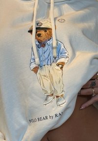 Sweatshirt branca com uma ilustração de um urso vestido com uma camisa riscada azul, calças bege e ténis brancos; texto "POLO BEAR by RAL" em baixo.