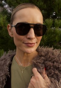 Lunettes de soleil noires surdimensionnées avec une finition brillante, portées avec des colliers superposés en argent et or, et un manteau en fourrure marron texturé. Haut vert visible.