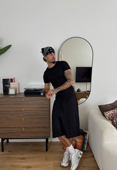 Camiseta negra y pantalones cortos en una persona, con tatuajes visibles. Una bandana negra, zapatillas blancas. Espejo redondo y cómoda de madera con decoración.