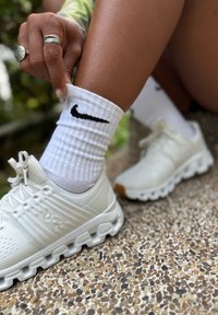 Sapatilhas brancas de desporto com parte superior em malha texturada e sola de borracha. Meias brancas com um logo preto da Nike. Mão a puxar a meia para baixo. Fundo em pedra.