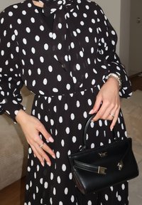 Vestido preto com grandes bolinhas brancas, com decote em laço e mangas bufantes longas. Segurando uma pequena bolsa preta com uma alça curvada.