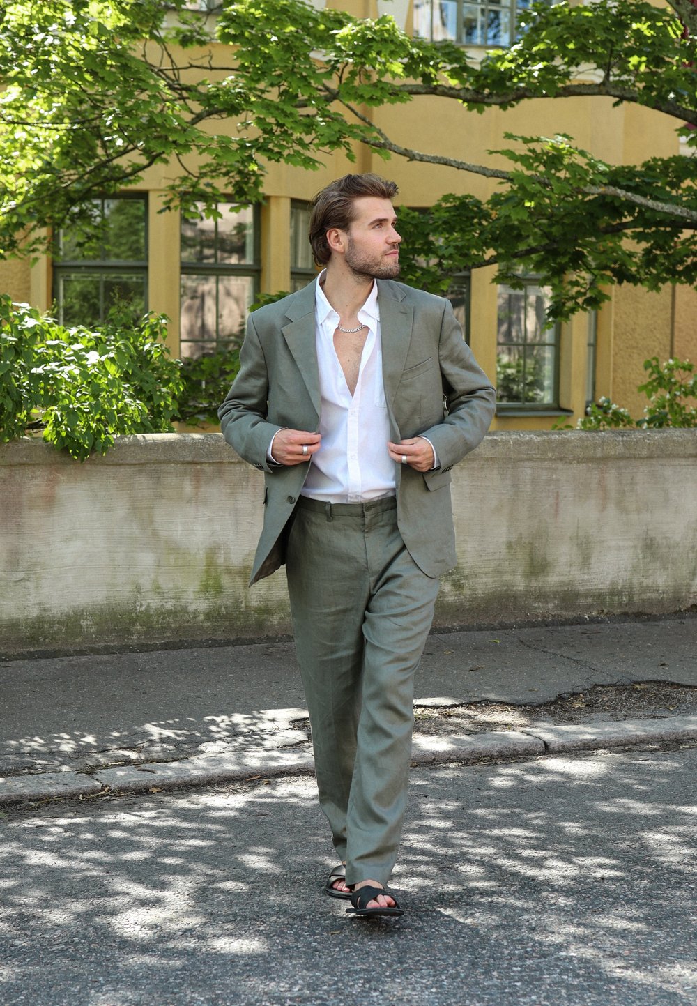 Traje de lino verde claro con solapa muesca, combinado con una camisa blanca desabotonada. Sandalias negras completan el look. Fondo natural.