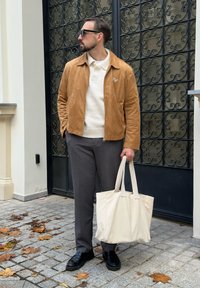 Férfi barna velúr dzsekiben, fehér pulóverben, szürke nadrágban, fekete cipőben, napszemüvegben, bézs táskát tartva, kövezett úton állva, fémból készült kapu háttérrel.