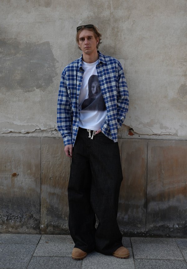 Joven con una camisa azul de cuadros sobre una camiseta blanca con gráfico, pantalones negros de pierna ancha y zapatos marrones, de pie contra una pared desgastada.