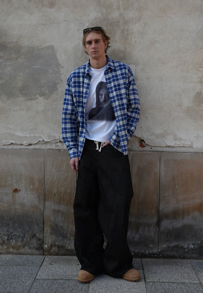Jeune homme portant une chemise à carreaux bleue par-dessus un t-shirt blanc avec un graphisme, un pantalon large noir et des chaussures marron, debout contre un mur usé.