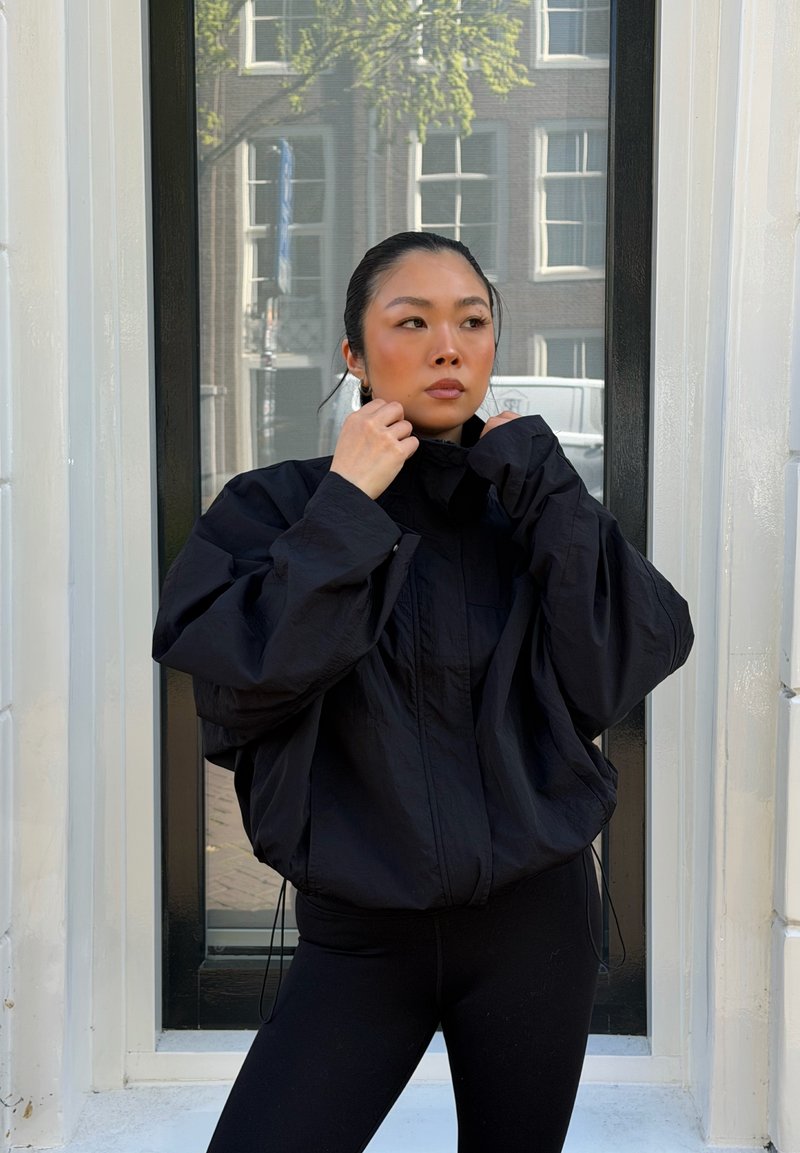 Femme ajuste le col d’une grande veste noire, debout devant une grande fenêtre avec le reflet d’un bâtiment.