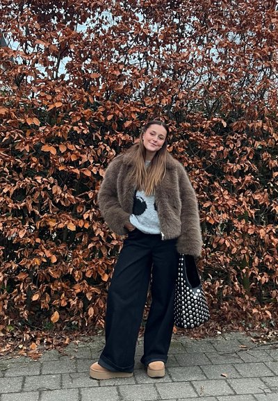 Mujer joven de pie al aire libre con pantalones anchos negros, zapatos beiges, suéter gris y chaqueta de piel marrón, sosteniendo un bolso negro con tachuelas junto a un seto de hojas marrones.