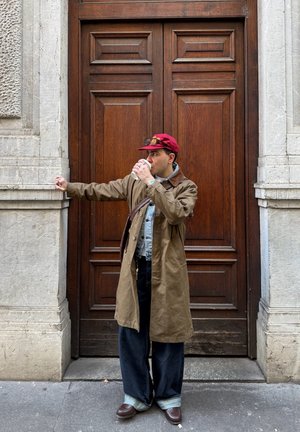 Hombre con una gorra roja y un abrigo marrón largo está apoyado contra una puerta de madera, bebiendo de una taza blanca, con un brazo apoyado en una pared de piedra.