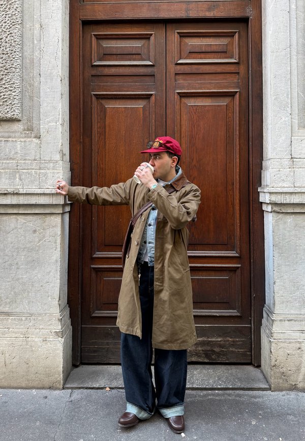 Man draagt een rode pet en een lange bruine jas, staat tegen een houten deur, drinkt uit een witte mok en leunt met één arm tegen een stenen muur.