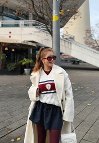 Palton alb din lână, pulover cu dungi roșii și albe, fustă bleumarin cu dungi fine, colanți vișinii și geantă albă; accesorizat cu ochelari de soare mari.