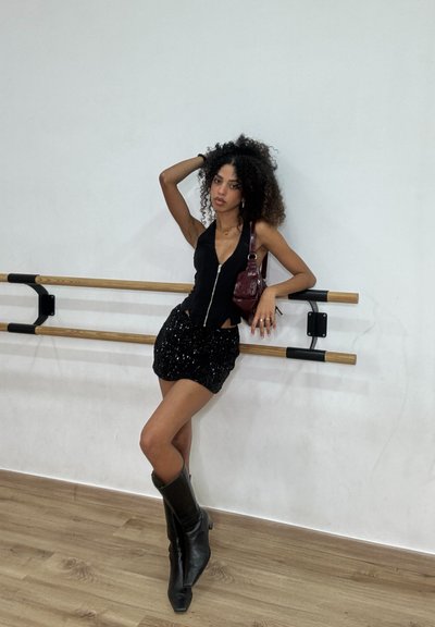 Body negro con cremallera combinado con una falda mini de lentejuelas. La modelo lleva botines negros brillantes hasta la rodilla y sostiene un pequeño bolso burdeos.