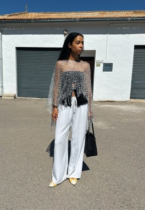 Top de flecos plateados y transparentes sobre una camiseta negra, combinado con pantalones blancos fluidos y tacones amarillos de punta. Sujetando un bolso negro.