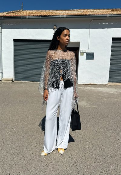 Top de flecos plateados y transparentes sobre una camiseta negra, combinado con pantalones blancos fluidos y tacones amarillos de punta. Sujetando un bolso negro.