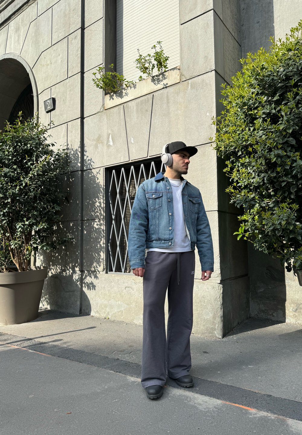 Chaqueta de mezclilla, camiseta blanca, pantalones anchos grises y calzado negro. Fondo de edificio gris con vegetación y una ventana.
