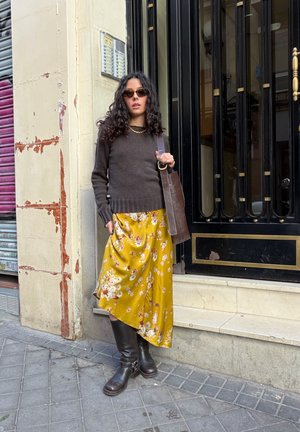 Suéter marrón, falda de satén amarilla con flores, botas marrones de cuero hasta la rodilla y bolso marrón. Cabello rizado, gafas de sol, de pie contra una pared.