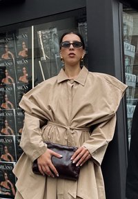 Palton oversized bej cu talie strânsă, asortat cu o poșetă de mână maro închis. Prezintă cercei mari aurii și ochelari de soare negri mici.