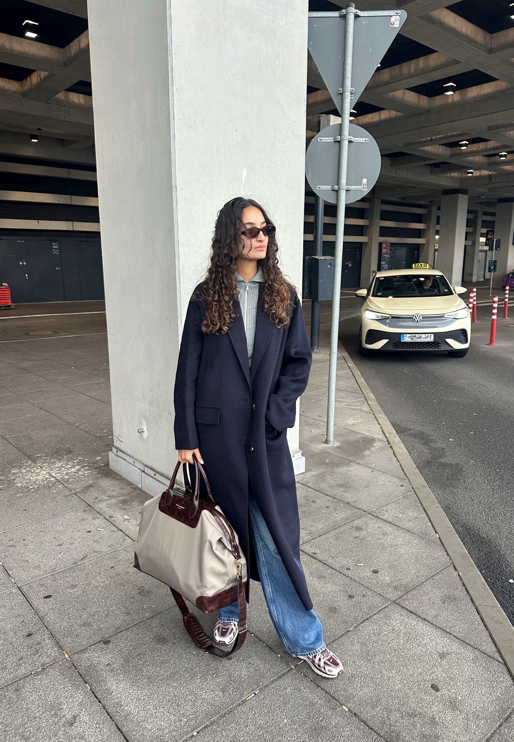 Casaco de lã azul-marinho com lapelas verticais, suéter cinza com textura, jeans de perna larga azul-claro e uma bolsa bege e bordô, acompanhados de ténis com padrão.