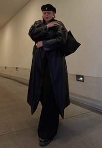 Μαύρο δερμάτινο μακρύ παλτό, φαρδιές μαύρες παντελόνες, μαύρο μπερέ και εντυπωσιακοί δακτύλιοι. Λείος υφασματικός πυρήνας με σύγχρονη σιλουέτα.