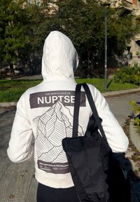 Hanorac alb cu un grafic de munți pe spate, care include textul "THE NORTH FACE" și "NUPTSE"; asortat cu o geantă de umăr neagră, purtată casual.