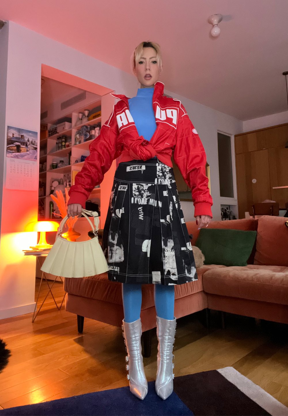 Veste rouge avec logo nouée à la taille, col roulé bleu, jupe plissée noire avec imprimé graphique, collants bleu clair, bottes argentées montantes jusqu'aux genoux, sac à main beige.