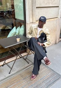 Pessoa sentada numa mesa de café, vestindo um casaco bege claro, camisa branca, calças pretas e sapatos castanhos. Chávenas de café na mesa.
