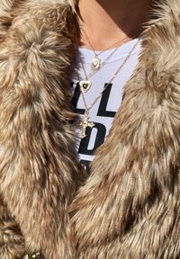 Faux fur frakke i lys brune nuancer med en blød, fluffy tekstur. Tre guldhalskæder lagdelt: et hjertevedhæng, en rektangulær ramme og et dekorativt kors.