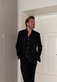 Homme en costume noir et chemise, appuyé contre l'encadrement d'une porte, les mains dans les poches, souriant et regardant sur le côté dans un environnement intérieur.