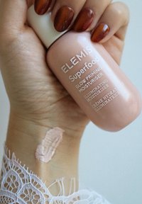 Crema idratante primer Elemis Superfood Glow in una bottiglia rosa tenuta in una mano con unghie marrone scuro; una piccola quantità di crema visibile sulla pelle.