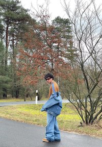 Giubbotto di denim sopra una camicia marrone, jeans larghi e strappati, e scarpe color sabbia. Ambientazione all'aperto con alberi e una strada asfaltata.