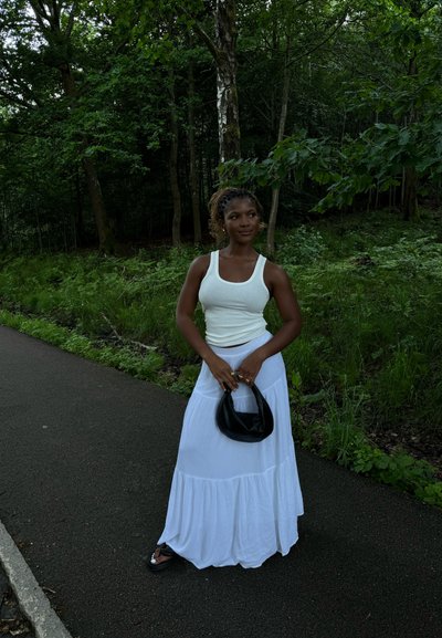 Top blanco sin mangas y falda blanca larga, sosteniendo un pequeño bolso negro. El conjunto presenta una textura suave y un dobladillo en capas. Fondo verde natural.