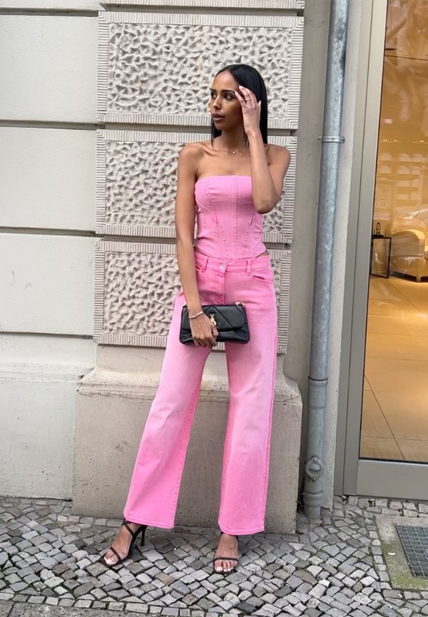 Haut en denim rose sans bretelles associé à un jean large assorti. Le mannequin tient une pochette noire et porte des sandales à talons noires. Mur texturé en arrière-plan.