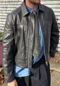 Zwarte leren jas met een voorrits, zijzakken en een gladde textuur, gedragen over een denim shirt en een groengekleurde stropdas met patroon.