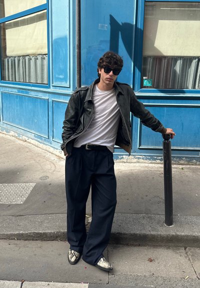 Chaqueta de cuero negra sobre una camiseta blanca, combinada con pantalones anchos color marino y zapatos estampados. Frente a una pared azul. Ambiente urbano casual.