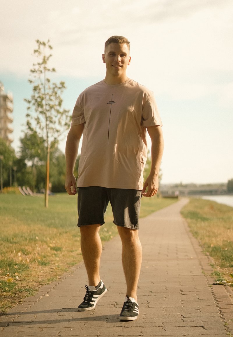 Jeune homme portant un t-shirt beige et un short foncé marchant sur un chemin pavé dans un parc près d'une rivière par une journée ensoleillée.