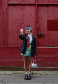 Veste en cuir noir sur une chemise blanche et une cravate en tartan rouge, jupe tie-dye pastel, tenant une tasse de café, avec un sac à main en argent et des chaussures à plateforme.