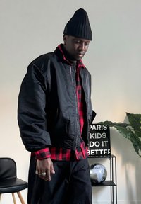 Zwarte bomberjack met een rood geruit shirt eronder, staande figuur, zwarte beanie, donkere broek, binnenomgeving met planten en decor.
