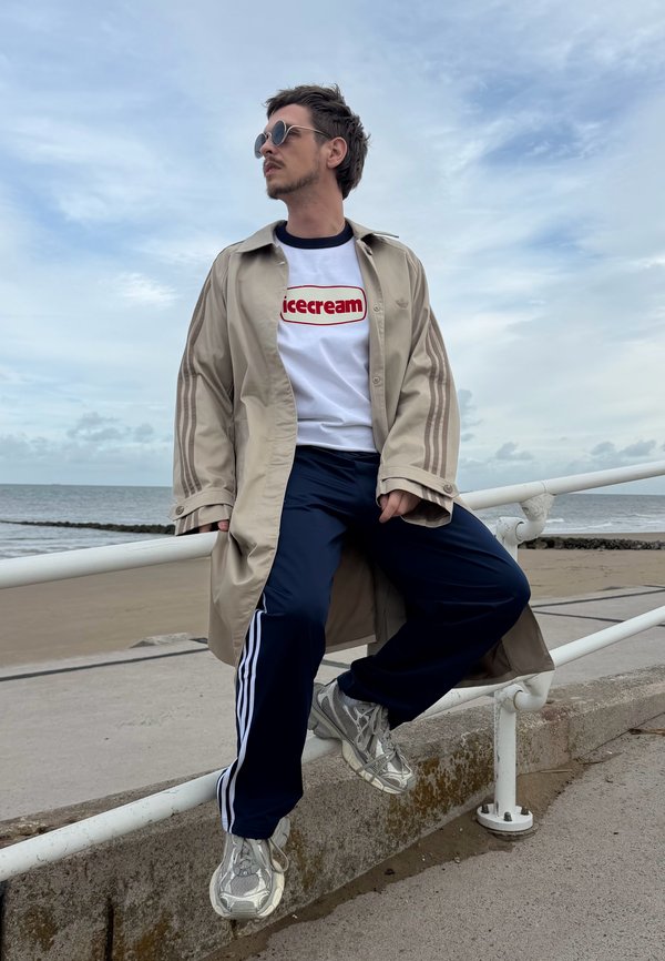 Hombre con gafas de sol, abrigo beige, camisa blanca "icecream" y pantalones deportivos azul marino sentado en una barandilla blanca junto a una playa bajo un cielo nublado.