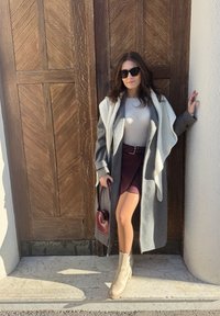 Manteau en laine gris avec col châle, haut blanc, jupe bordeaux à fines rayures, bottines beige, et sac à main rouge en forme de cœur devant une porte en bois.